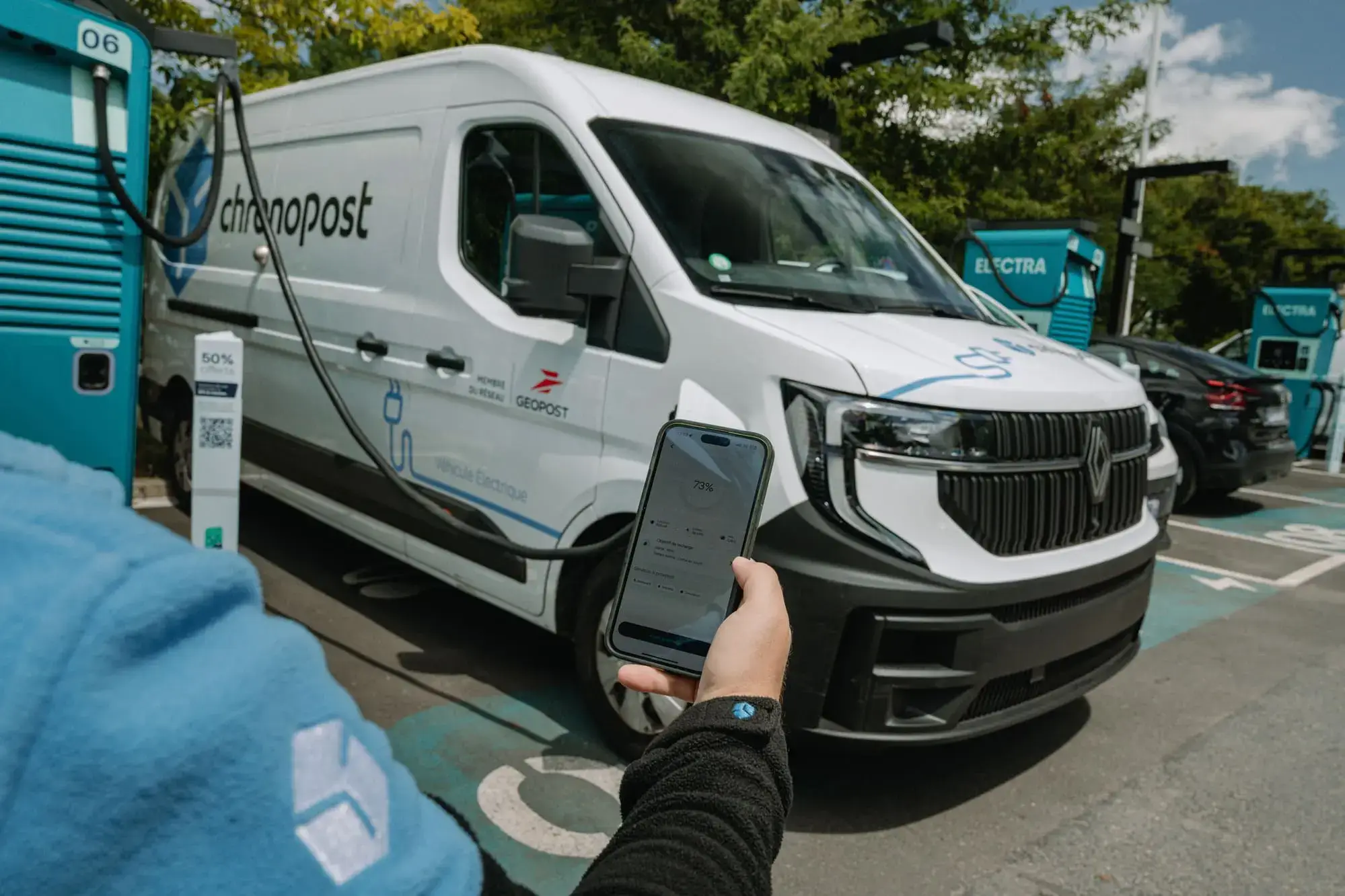Person holding a smartphone in front of a white delivery van parked at an Electra charging station, surrounded by trees.