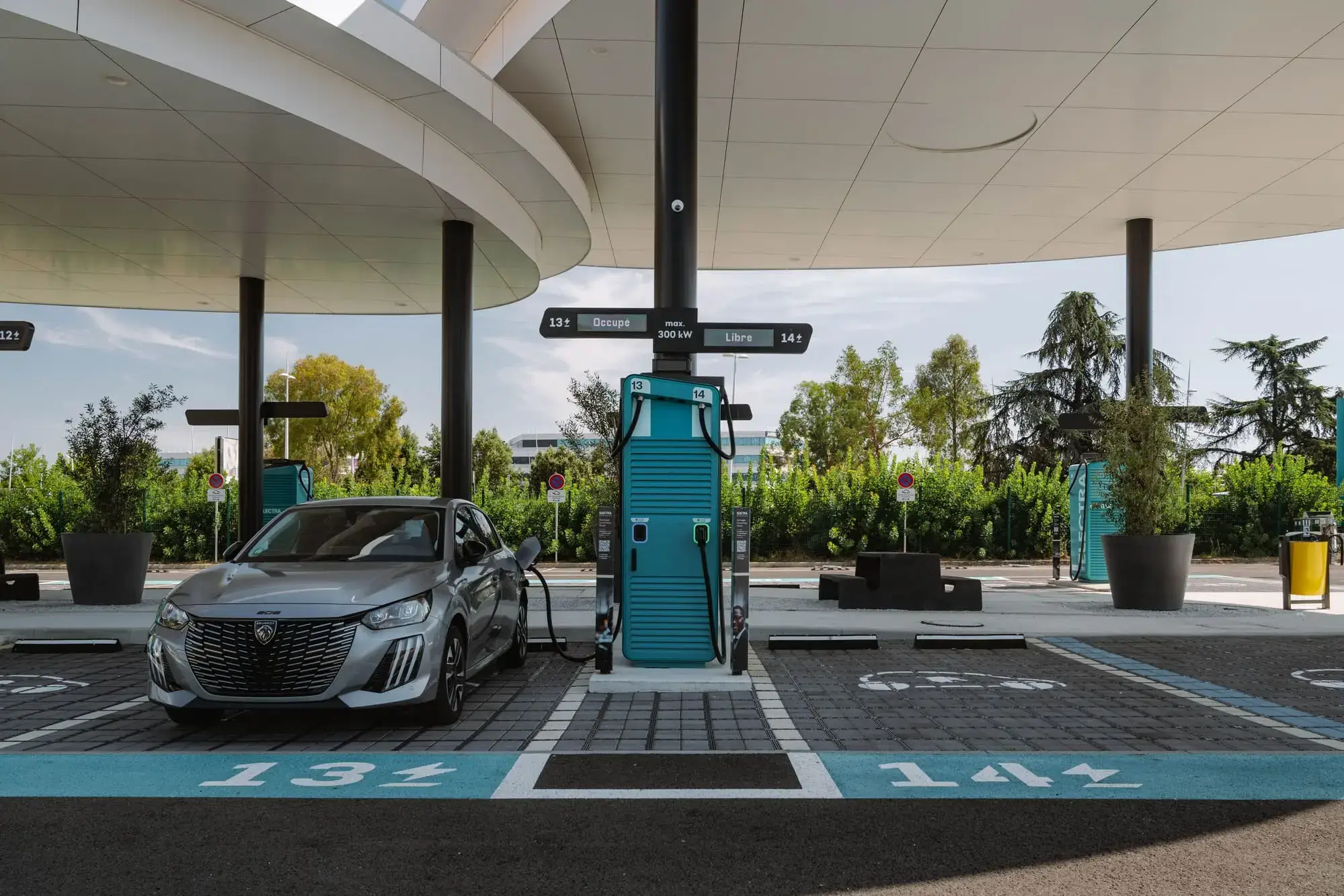 Electric car charging at a modern Electra station with multiple charging points, surrounded by greenery and a clear sky.