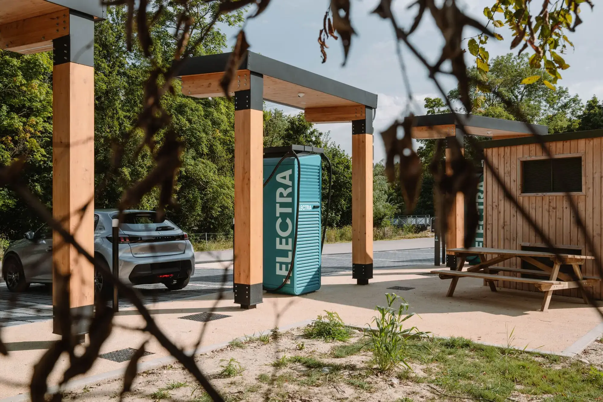 Electric car charging at an Electra charging station with wooden structures, surrounded by greenery and a picnic table in the foreground.