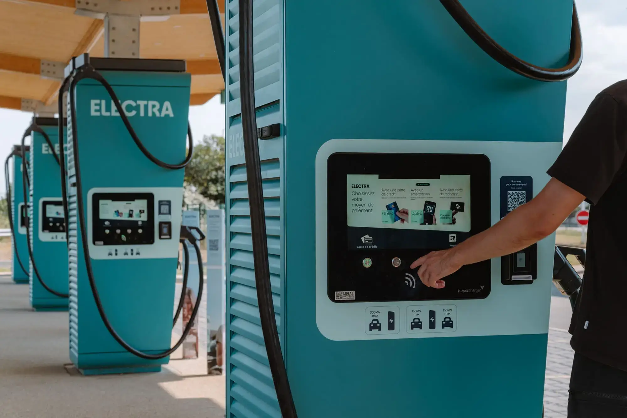 Person using a touchscreen on a teal electric vehicle charging station labeled "ELECTRA" at an outdoor charging area.