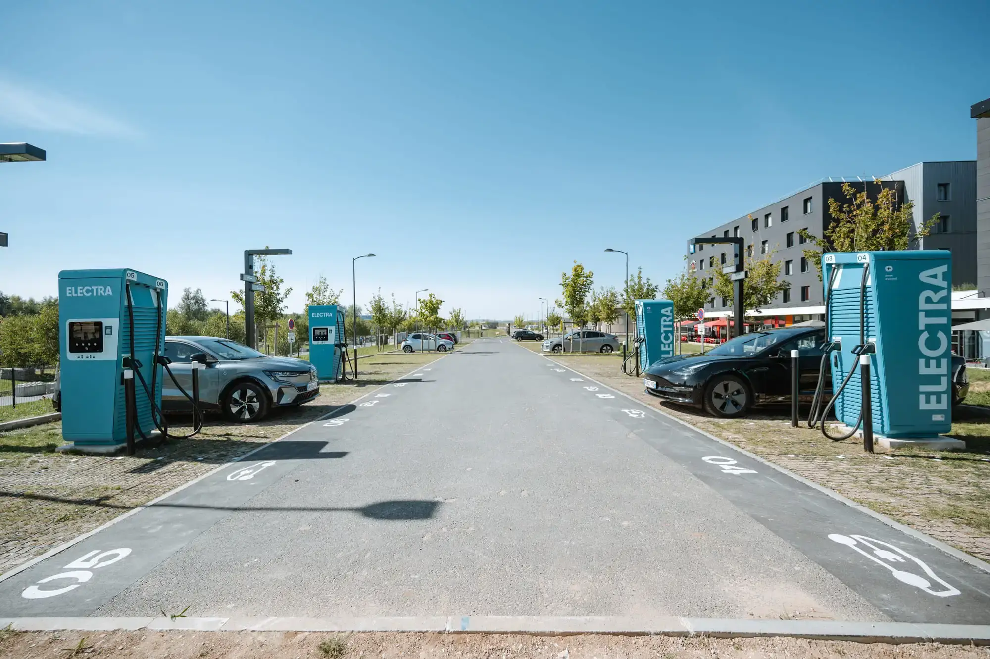 Electric car charging station by Electra with two cars plugged in, surrounded by trees and modern buildings under a clear blue sky.