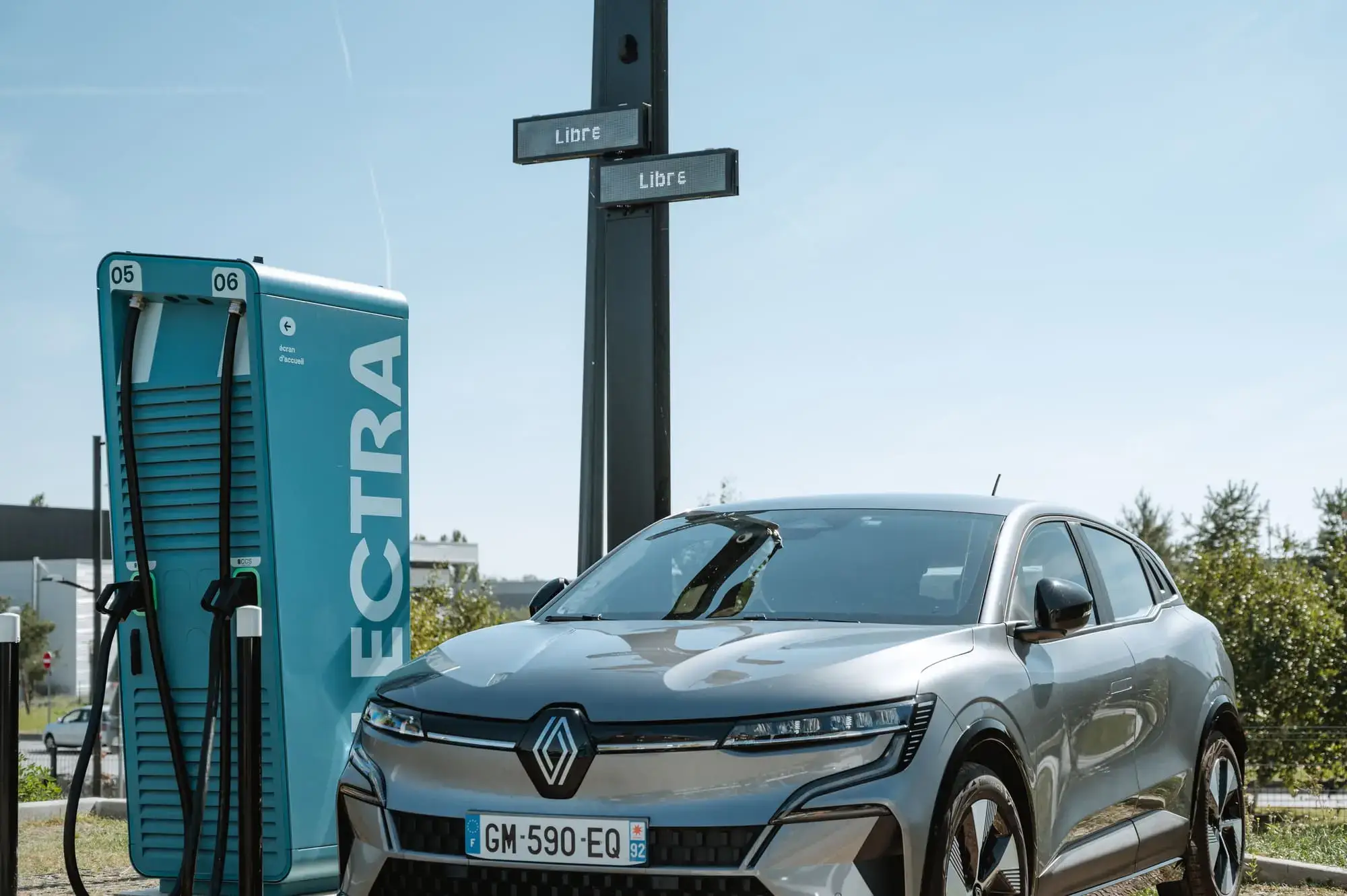 Silver electric car charging at a blue Electra station under a clear sky, with trees and a "Libre" sign in the background.