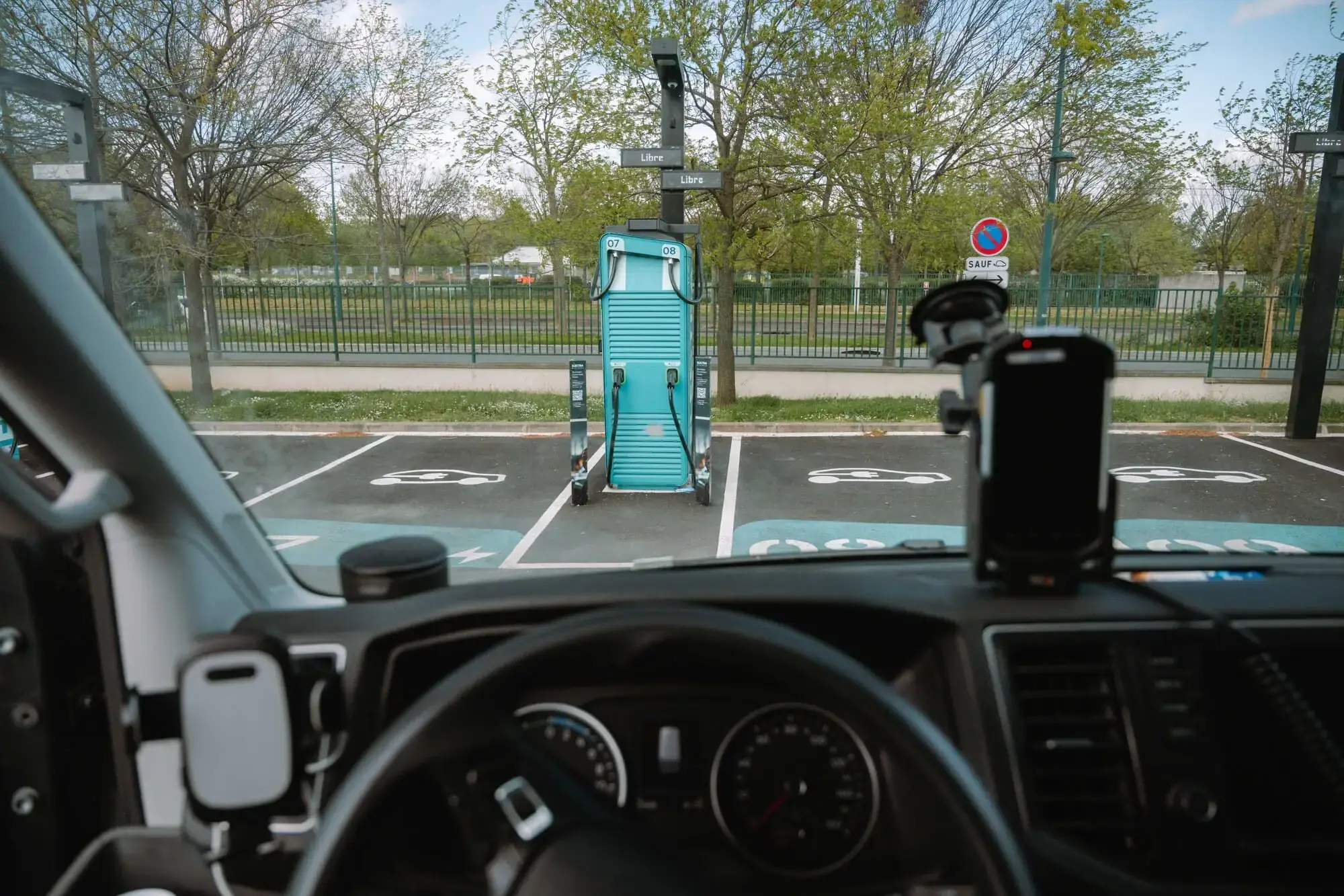 View from inside a vehicle showing an electric vehicle charging at an Electra station in a parking lot with trees and a fence in the background.