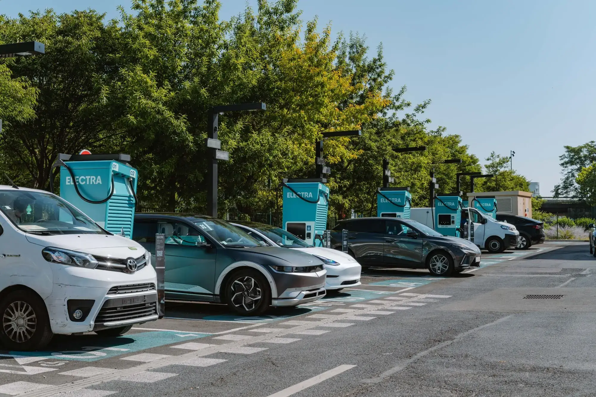 Electric vehicles charging at a row of charging stations in an Electra parking lot, surrounded by trees under a clear blue sky.