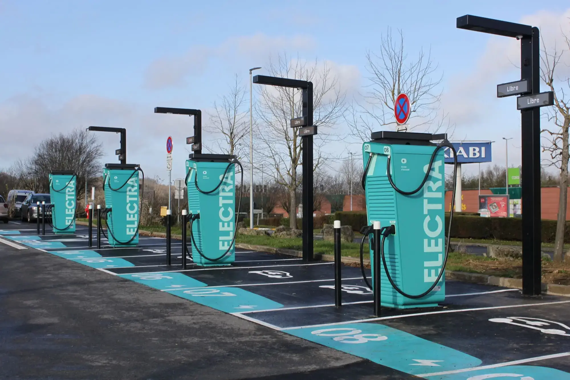 Four electric vehicle at an ELECTRA branded charging stations with teal branding in a parking lot, surrounded by trees and a store sign in the background.