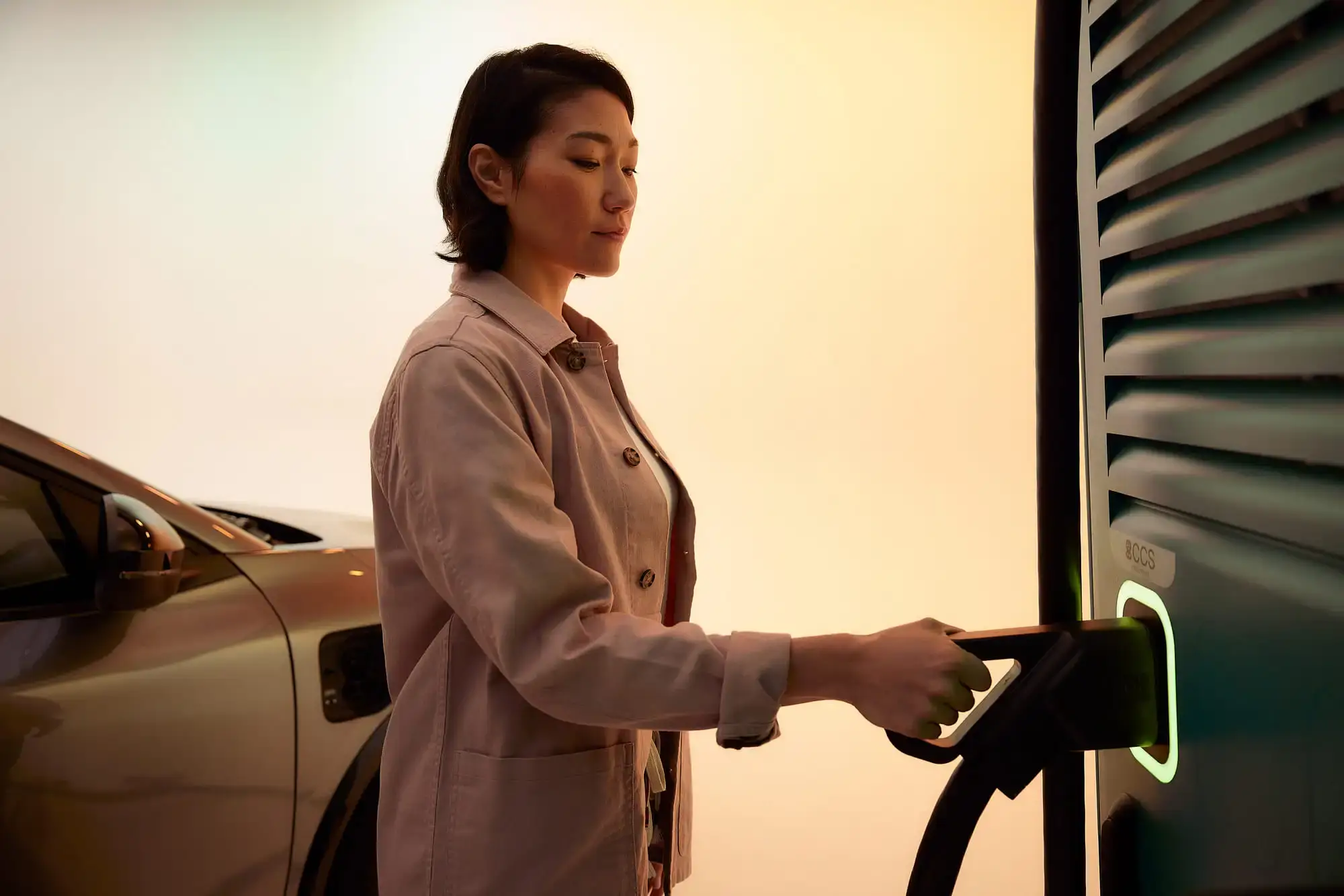 A person plugs a charging cable into an electric vehicle at an Electra charging station, with a warm-toned background.