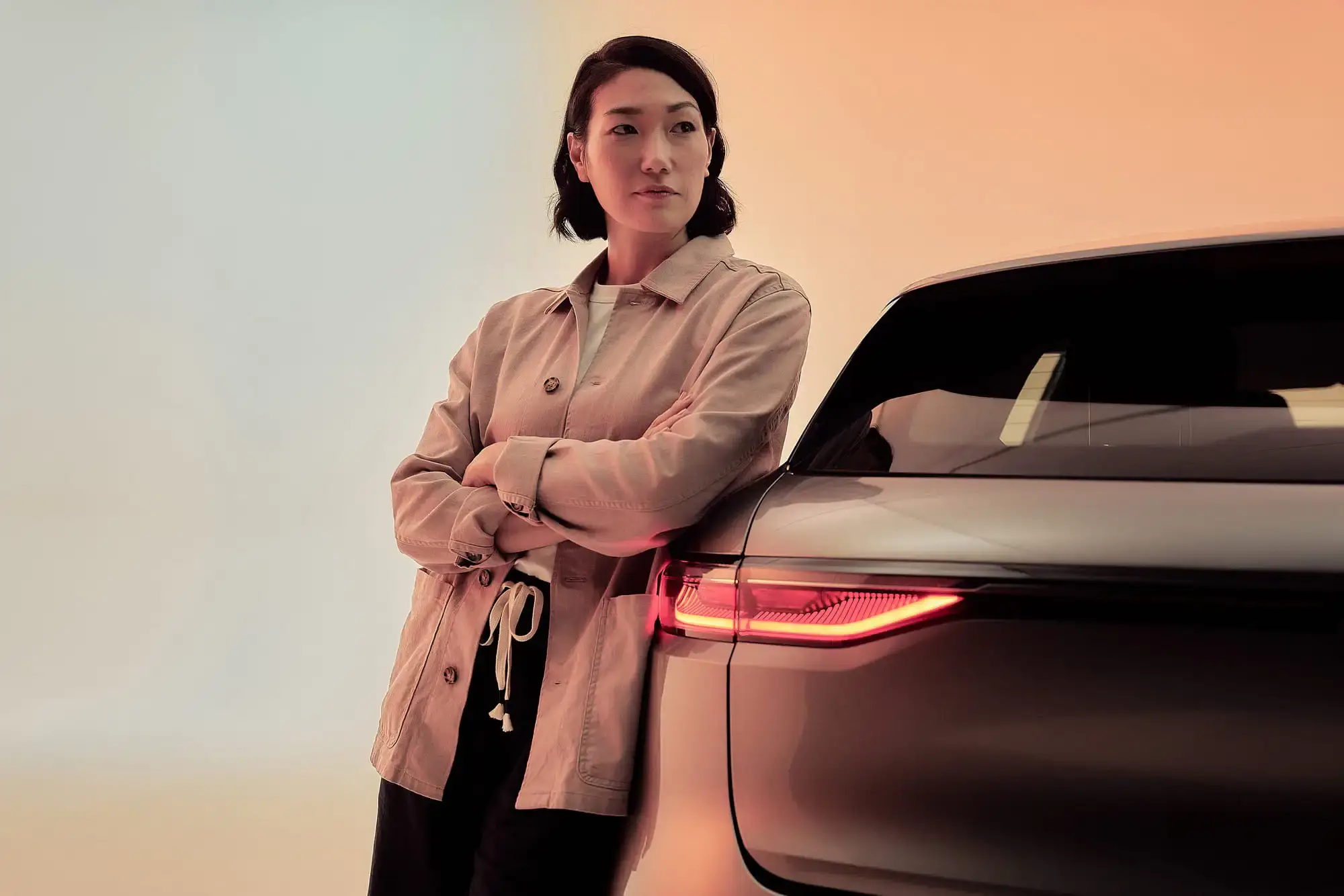 Woman in a beige jacket leans against a sleek black car with illuminated taillights, set against a gradient background for an ELECTRA VISUAL ASSET